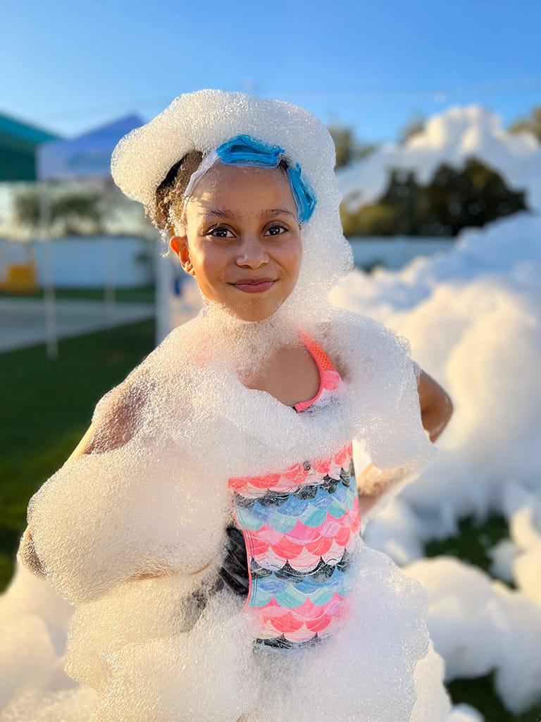 A girl wrapped shoulder-to-head in fresh foam, grinning at the camera