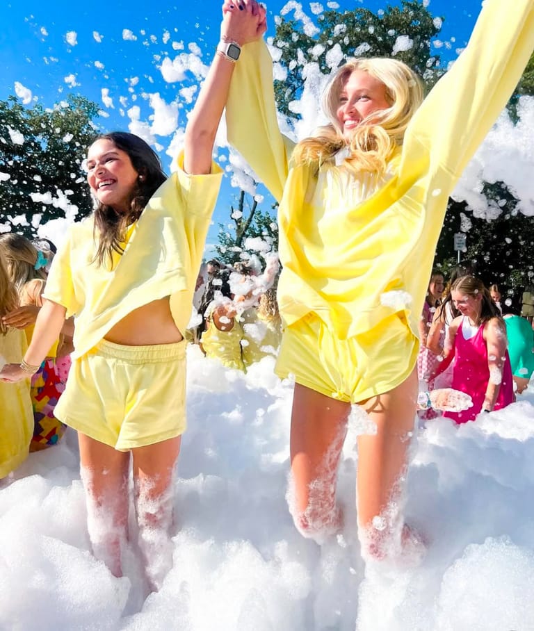 Two girls in matching yellow outfits holding hands overhead as they wade through knee-deep foam