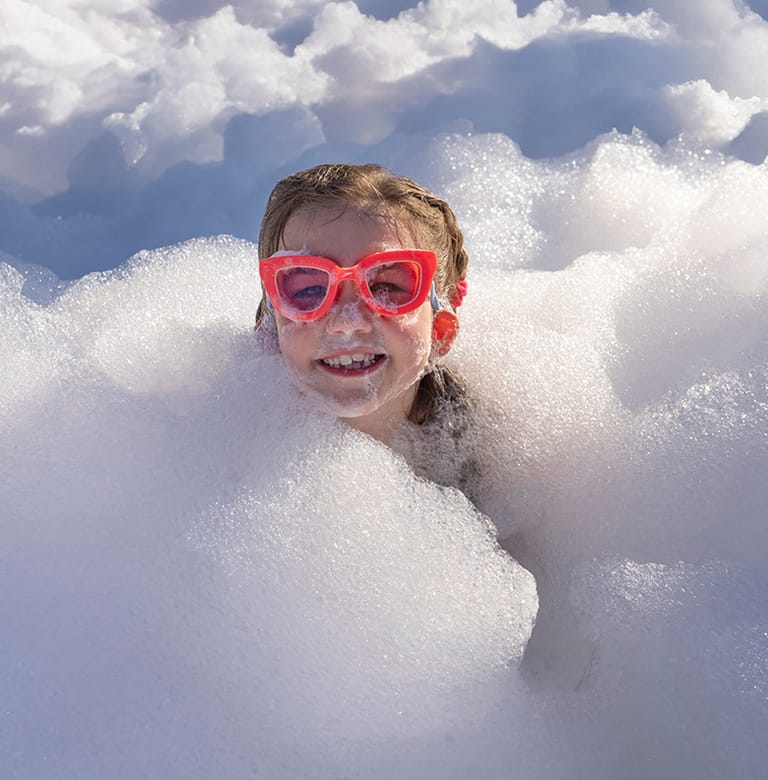 A smiling kid in swim goggles chest-deep in a drift of white foam