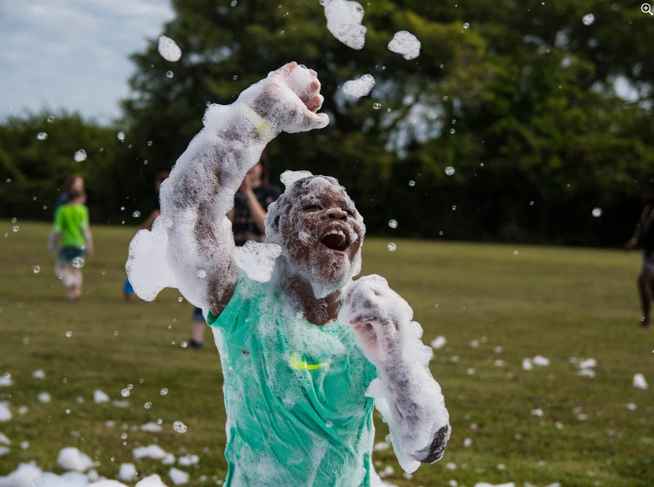 A kid mid-shout with an arm in the air, covered in foam on an open lawn