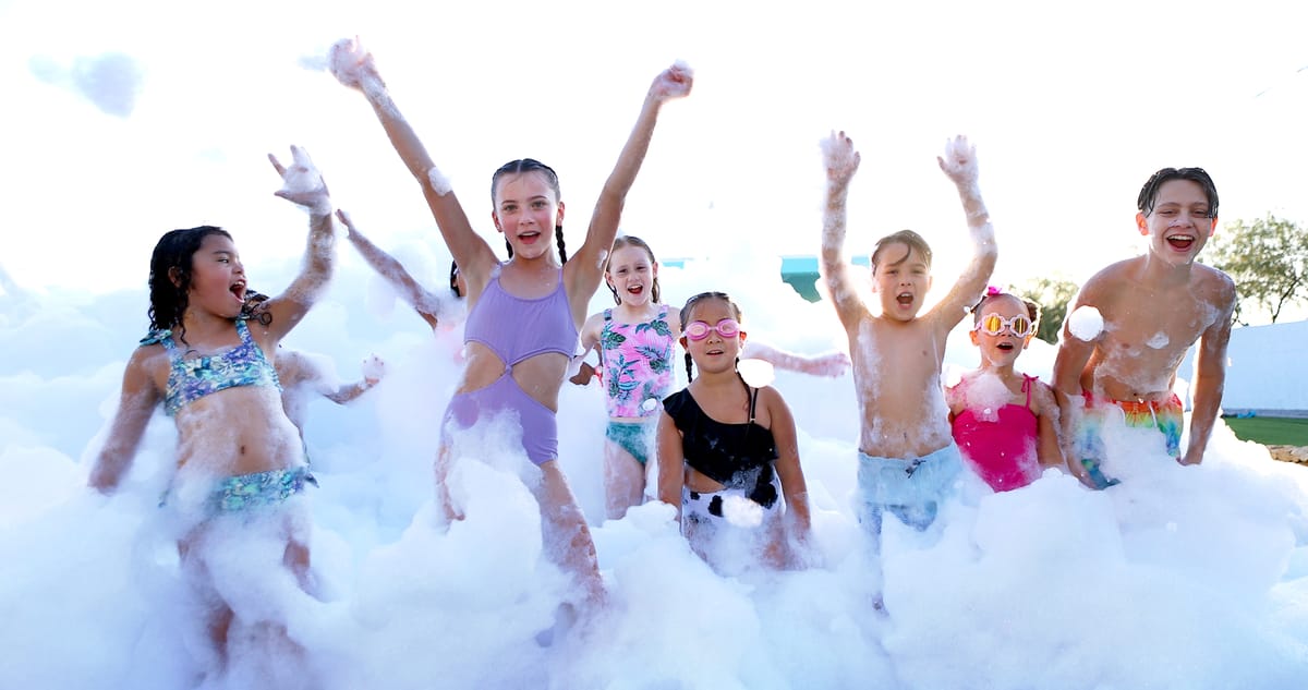 Seven kids standing waist-deep in foam with their arms raised, cheering