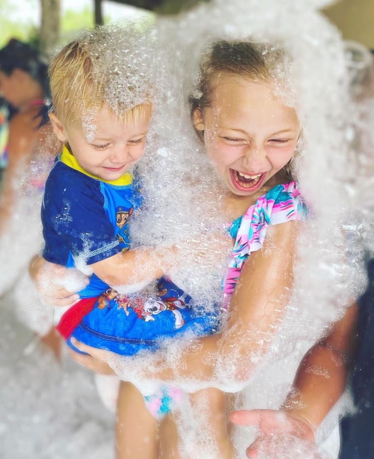 A girl carrying a toddler through deep foam, both of them laughing with their eyes squeezed shut