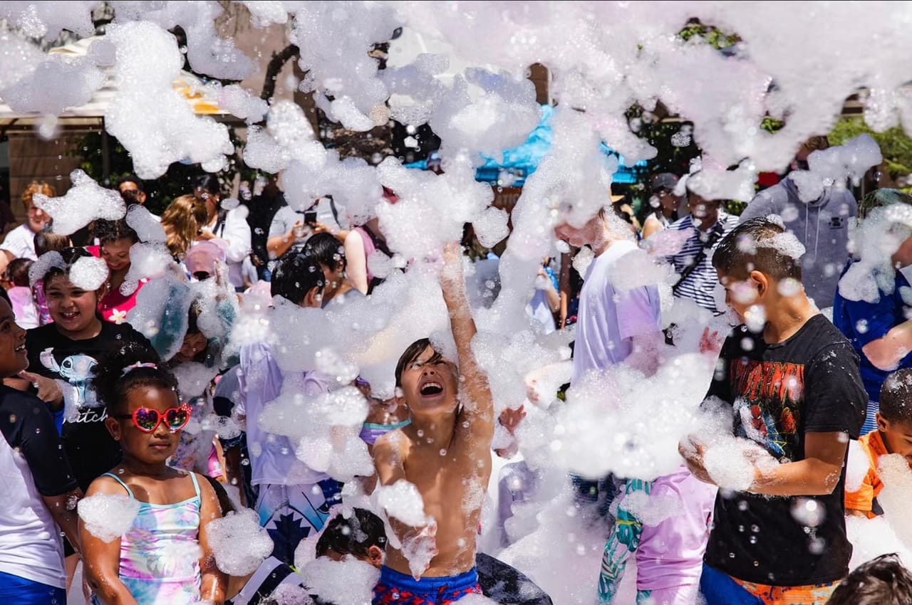 A crowd of kids outside under a cloud of foam, one of them laughing with an arm up to catch it